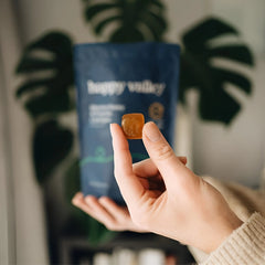 Happy Valley Honey Manuka Honey & Propolis lozenge held by fingers, with blue pouch in background.